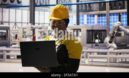 Ritratto di un ingegnere sorridente di una fabbrica di pannelli solari che analizza i dati su un laptop per prevedere guasti agli ingranaggi. Felice dipendente di un impianto fotovoltaico che utilizza un taccuino, seguendo le procedure di manutenzione, fotocamera A Foto Stock