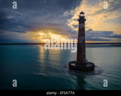 I raggi del sole si riversano da dietro una banchina di nuvole mentre il faro di Morris Island, ora fuori uso, sorveglia l’ingresso del porto di Charleston. Il faro Foto Stock