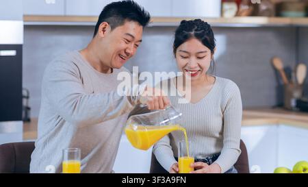 Uomo asiatico felice che versa succo fresco per una donna asiatica sorridente in una cucina moderna, concetto di stile di vita sano Foto Stock