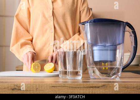 Donna che taglia un limone fresco su un tagliere bianco accanto a una brocca filtrante e due bicchieri d'acqua, Scelte di stile di vita sano, Idratazione Foto Stock
