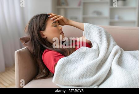Donna asiatica esausta che tocca la fronte, sdraiata sul divano sotto la coperta Foto Stock