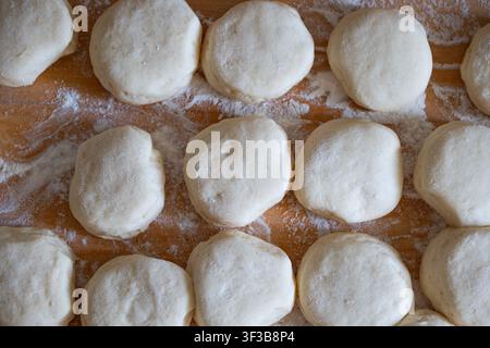 Una vista dall'alto ad angolo elevato che mostra file di porzioni di impasto lievitato rotondo e crudo appena tagliate, spolverate di farina su una superficie di tavola da pasticceria in legno, preparate per Baki Foto Stock