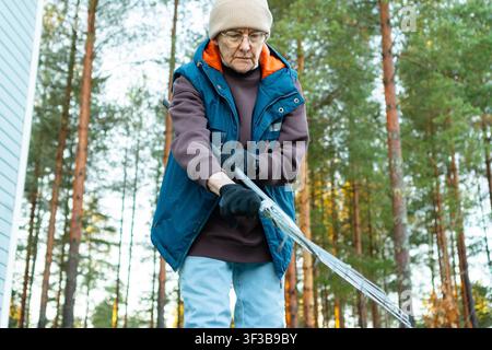 Donna anziana serena che apre un rastrello a ventaglio per il lavoro in giardino nella foresta autunnale. Anziana resiliente mostra indipendenza e mobilità nonostante l'età. Pensionata attiva gode dei compiti all'aperto in una giornata gelida tra gli alberi Foto Stock