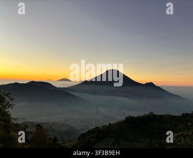 Stratovulcano che si erge sopra una valle piena di nebbia all'alba, con una piccola città visibile sotto e picchi stratificati che svaniscono nell'orizzonte dorato. Foto Stock