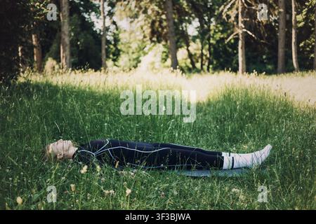 Donna in abbigliamento sportivo sdraiata su un materassino tra l'erba alta nel parco. Pratica di restaurazione dello yoga nidra, rilassamento profondo, meditazione per il sonno all'aperto. Foto Stock
