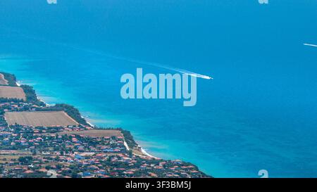 Ripresa a droni che mostra una zona residenziale costiera con campi, un mare turchese limpido e un motoscafo che lascia una scia bianca. Foto Stock