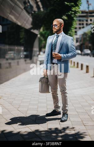 Uomo d'affari sicuro di sé in giacca blu che tiene un caffè e una valigetta camminando sul marciapiede della città Foto Stock