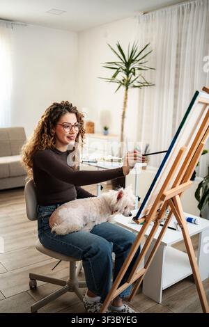 Giovane donna adulta che dipinge su cavalletto con un cane nello studio di casa Foto Stock