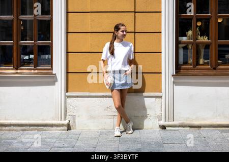 Giovane donna in piedi davanti a un edificio giallo illuminato dal sole. Giovane donna in abiti estivi casual in piedi davanti a un edificio giallo illuminato dal sole tra due Foto Stock