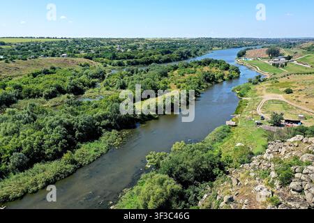 Un fiume scorre attraverso campi verdi e zone boschive sotto un cielo blu terso. Il paesaggio presenta colline sullo sfondo con un sentiero sterrato lungo il Foto Stock