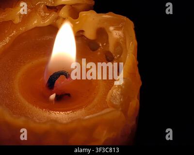 Vista macro di una singola candela di cera d'api accesa con una fiamma arancione vivace, che mostra texture dettagliate di bordi di cera dorata fusa su uno sfondo nero Foto Stock