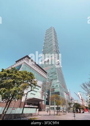 Taipei 101 si erge drammaticamente contro un cielo blu terso in una vista a bassa angolazione dal distretto di Xinyi, Taipei. L'iconica torre a gradini in vetro verde domina lo skyline sopra gli alberi e i blocchi urbani circostanti. Foto Stock