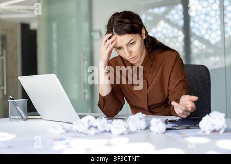 Donna che si sente sopraffatta, si tiene la testa mentre lavora su un laptop con carte accartocciate sparse sulla scrivania, simboleggiando frustrazione lavorativa e una scadenza difficile Foto Stock