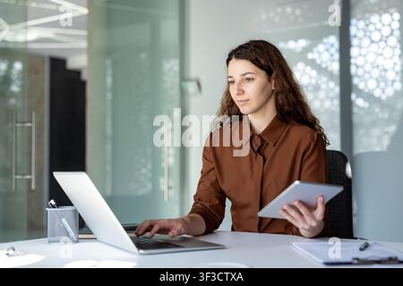 Giovane donna d'affari che lavora in modo efficiente in un ufficio moderno, bilanciando compiti sul suo laptop e un tablet digitale, incarnando produttività e dedizione professionale nel suo lavoro quotidiano Foto Stock