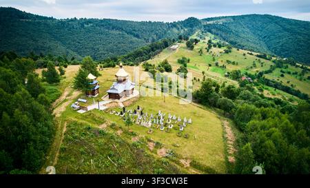 Paesaggio aereo con cappella di legno e cimitero di villaggio su una collina erbosa nei Carpazi. Utile per la pubblicità turistica, il patrimonio rurale, la spiritualità e i concetti di natura Foto Stock