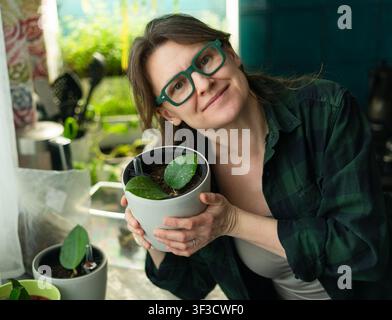 Una donna gioiosa tiene un seme di pianta in vaso e sorride in una stanza soleggiata a casa. La felice genitore di piante culla la vegetazione indoor e il germoglio di pianta d'appartamento con orgoglio caloroso vicino a una finestra luminosa. Foto Stock