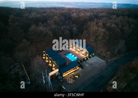 Vista aerea notturna mostra cabine moderne del resort intorno a una piscina luminosa. Luci calde, terrazza in legno spaziosa e ambientazione boschiva isolata creano un concetto di ritiro di lusso invitante Foto Stock