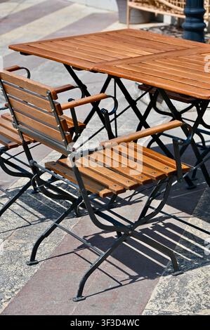 Tavoli pieghevoli in legno vuoti e sedie disposti su una terrazza esterna soleggiata lastricata con piastrelle di pietra, pronti per gli ospiti di un caffè o ristorante di strada mediterraneo. Foto Stock