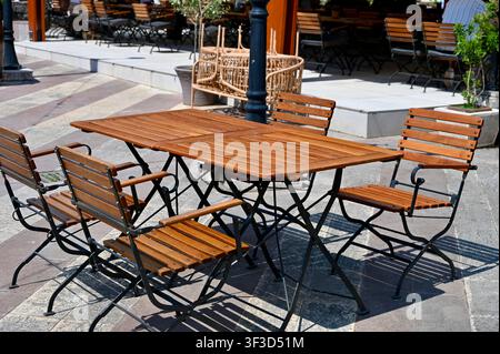 Tavoli pieghevoli in legno vuoti e sedie disposti su una terrazza esterna soleggiata lastricata con piastrelle di pietra, pronti per gli ospiti di un caffè o ristorante di strada mediterraneo. Foto Stock