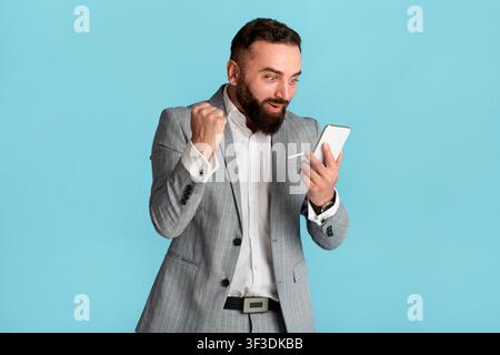 Eccitato giovane uomo d'affari guardando smartphone e facendo sì gesto su sfondo blu Foto Stock