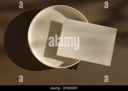 Modello di biglietto di auguri vuoto minimale su piatto di ceramica su sfondo di tavolo marrone. Luce naturale calda, ombre dure. Modello elegante di cartoleria estiva Foto Stock