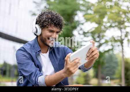 Giovane uomo immerso nel gioco digitale su un tablet mentre indossa cuffie bianche, sorridendo ed esprimendo entusiasmo durante il suo tempo libero in un ambiente all'aperto Foto Stock