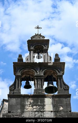Vista ad angolo basso di un vecchio campanile in pietra con campane di bronzo e una croce di metallo in cima, sullo sfondo un cielo blu nuvoloso. Ideale per concetti di religione, spiritualità, storia, eredità cristiana e architettura europea. Foto Stock