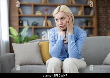 Donna matura seduta su un comodo divano grigio nel suo salotto, appoggiando le mani sulle guance, sembrando pensierosa e riflettendo su sfide personali o stress emotivo Foto Stock