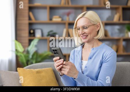 Donna anziana che si rilassa su un divano, si impegna felicemente con il suo telefono cellulare, si connette con gli altri e gode della comunicazione digitale in un ambiente confortevole del soggiorno Foto Stock