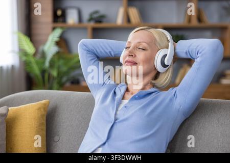 Donna anziana che si rilassa su un comodo divano a casa, chiudendo gli occhi felicemente mentre ascolta musica attraverso cuffie moderne, trovando pace e tranquillità durante il tempo libero Foto Stock