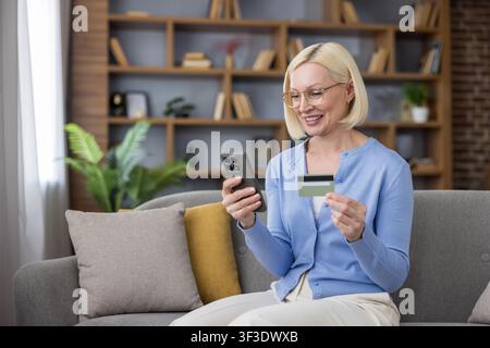Donna anziana seduta su un divano, sorridente e con in mano uno smartphone e una carta di credito, godendo dello shopping e delle operazioni bancarie online convenienti da casa Foto Stock
