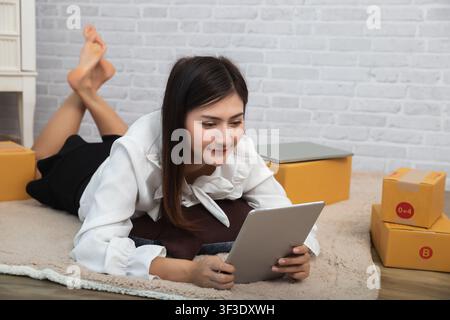 Donna d'affari accetta ordini dai clienti tramite vendite online mentre è sdraiata su un tappeto nel suo ufficio domestico, prepara una scatola di pacchi di prodotti per la consegna Foto Stock