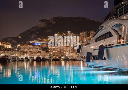 MONACO - 20 GIUGNO 2026: Superyacht di lusso moderna con luci LED blu subacquee ormeggiata al porto di Port Hercule di notte con lo skyline di Monte Carlo in b Foto Stock