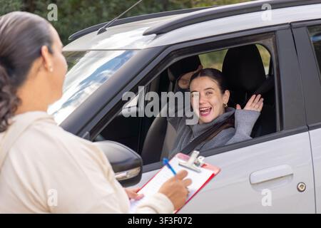 Giovane studentessa solleva le mani e ride allegramente mentre l'istruttore di guida scrive appunti su una clipboard, finestrino dell'auto, concetto di celebrazione Foto Stock