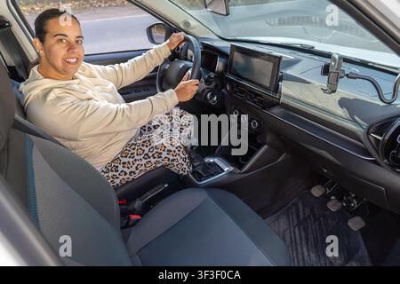 Donna sul sedile del conducente sorride e guarda verso la fotocamera, angolazione dall'alto, interno dell'auto con cruscotto e supporto per telefono visibile Foto Stock