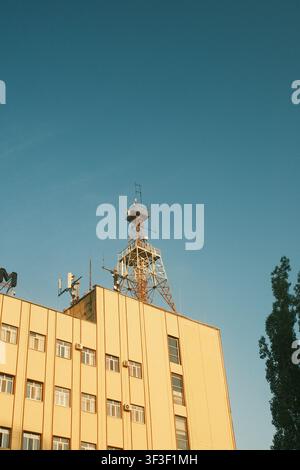 Torre radio per telecomunicazioni e antenne sulla sommità di un edificio industriale giallo contro un cielo blu chiaro durante l'ora dorata. Foto Stock