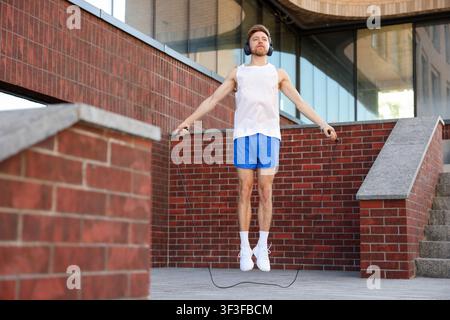 Uomo in abbigliamento sportivo e cuffie che fa esercizio con la corda per saltare vicino a un edificio all'aperto, vista dall'alto Foto Stock