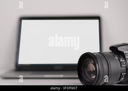 Primo piano di una fotocamera digitale e di un obiettivo accanto a un laptop con schermo bianco vuoto, un mockup di spazio di lavoro minimalista per fotografia, editing, portfolio e c Foto Stock
