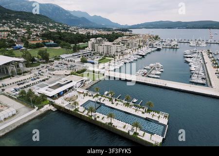 Vista aerea di una marina con yacht di lusso, incastonata contro lo sfondo montuoso, delimitata da una moderna passeggiata, parcheggio adiacente e hotel sul mare, blu chiaro Foto Stock