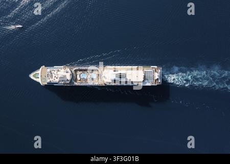Vista aerea di una nave da crociera che scivola sull'oceano blu profondo, ponti illuminati dal sole con piscina e scialuppe di salvataggio, scia sottile, simmetria panoramica, a bordo Foto Stock