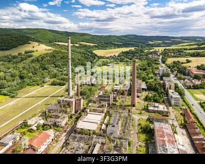 Veduta aerea dell'ex fabbrica chimica slovacca abbandonata con alti camini in mattoni, edifici industriali in rovina e vegetazione invasa in Foto Stock
