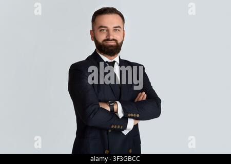 Bellissimo CEO barbuto in abbigliamento formale in piedi con braccia incrociate su sfondo grigio chiaro Foto Stock