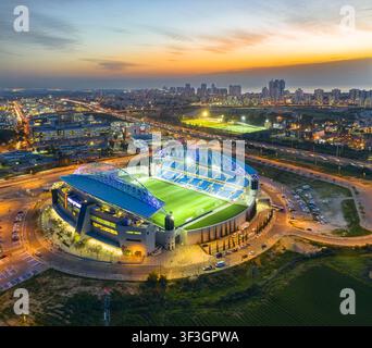 Vista aerea dello stadio di Netanya illuminato al tramonto con lo skyline della città sullo sfondo a Netanya, Distretto Centrale, Israele. Foto Stock