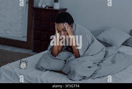Ragazza afroamericana in coperta siede a letto con la sveglia Foto Stock