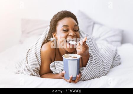 Gelato a letto. Donna afroamericana sorridente avvolta in coperta e mangiando Foto Stock