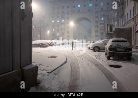 Una donna solitaria cammina di notte attraverso un cortile cittadino ricoperto di neve, circondata da auto parcheggiate e alti palazzi residenziali. Foto Stock