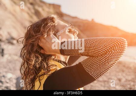 Donna spiaggia sole giovane donna con capelli ricci gode della luce calda del tramonto sulla riva rocciosa indossando occhiali da sole e giubbotto di salvataggio Foto Stock