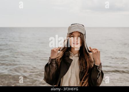 Cappello da spiaggia da donna giovane donna con giacca di pelle e caldo ushanka in riva al mare con spazio di copia per viaggi stagionali Foto Stock
