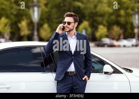 Maganer finanziario serio in abbigliamento formale che parla sul cellulare vicino alla sua auto a grande città Foto Stock