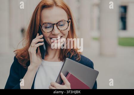 Donna d'affari dai capelli rossi con occhiali e giacca parla al telefono e tiene un tablet e un blocco appunti mentre cammina all'aperto vicino a un edificio per uffici. Foto Stock